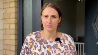 A woman, Kelly Heather, wearing a colourful top and standing in the doorway of her home