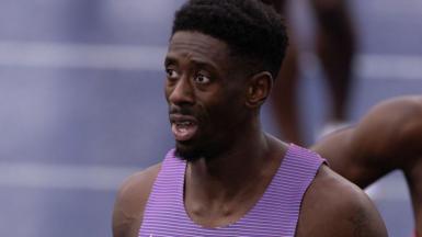 Close-up photograph of Reece Prescod following a 100m race. He has black hair and a goatee beard, and is wearing a purple running vest. Part of the track can be seen in the background.