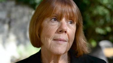 Gisèle Pelicot, a woman with chestnut hair in a bob, pictured in the dappled light of a garden or park, in close-up, looking up and to the left, with the background blurred behind her