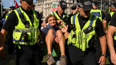 Three male police officers detain a female protester, who is holding a paper sign appearing to mention Palestine, during a rally challenging the UK government's proscription of "Palestine Action" under anti-terrorism law. In the background there are grand buildings a Palestine flag and a poster mentioning genocide