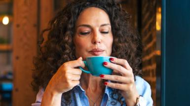 A woman with dark curly hair sits in a cafe and blows on her coffee to cool it down. She looks quietly happy. Her red nail polish stands out against the ocean blue of the coffee cup.