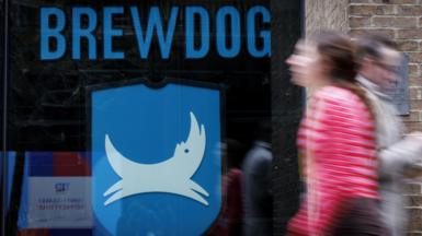 People walk past a closed-down BrewDog bar in central London. The window of the pub has the word Brewdog written in large blue letters above its logo, featuring a white dog on a blue background. 