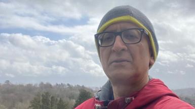 Dr Ravi Kumar - wearing black glasses, a green beanie and red jacket - taking a selfie with trees stretching into the horizon behind him