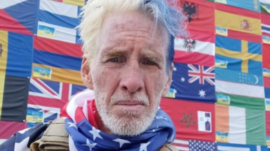 man stands shooting a selfie with blue and grey hair, a military vest and an American flag neck scarf. A large pattern of flags are behind him as a backdrop