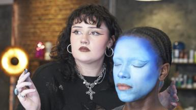 Ailish is standing next to a seated make-up model. Ailish is holding a thin brush in one hand and has two turquoise rings on her fingers. She is looking off to the side, probably at a mirror, rather than directly at the model and has a look of concentration on her face. Ailish has black wavy hair with a fringe and bold black eyeliner. She is also wearing a black distressed jumper, silver hoop earrings and a silver necklace with a cross on it. She also has three silver nose piercings, including a nose ring, stud and septum barbell. The model has her eyes closed and her entire face has been painted using different shades of blue. It is clear that the make-up look has not been complete yet and the model's lips are bare. The model's black hair has been plaited back into cornrows. The background is out of focus but Ailish and the model are in a make-up studio with shelves or products and a ring light behind them. 