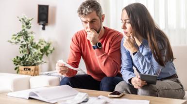Couple sit on a sofa looking at paperwork with worried looks on their faces.
