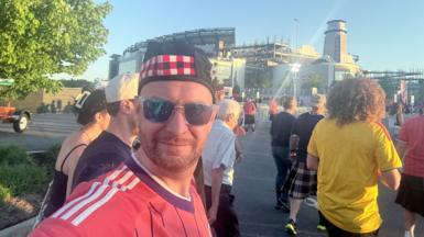 Man wears a Scotland top, hat and sunglasses, outside a sport stadium