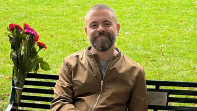 Geraint sitting on his wife's memorial bench He is in a a brown collarless jacket with a zip and khaki trousers. There is a bunch of flowers tied to the bench.