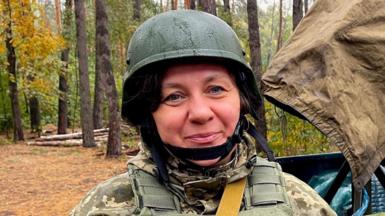 Inna stands in a forest wearing military fatigues and a helmet