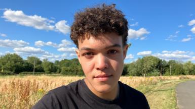 A young man with dark curly brown hair standing by the side of a road. He is wearing a black t-shirt and behind him are yellow crops and blue skies.