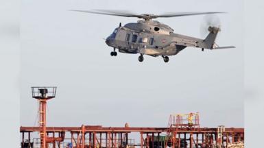 A grey helicopter seen above an orange and blue oil tanker