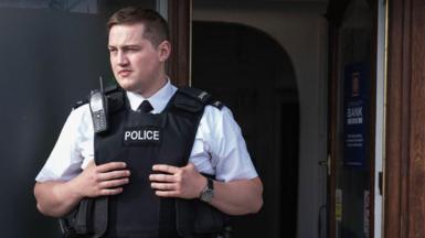 Finnian Garbutt, a man with dark hair, wearing a white shirt with black tie and a black support vest with the words police on it. He is wearing a black watch on his left wrist and there is a brown wooden door in the background.