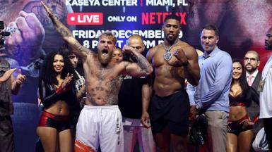 Jake Paul points his arm in the air as he and Anthony Joshua stand side by side at a weigh-in