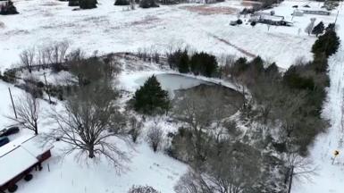 A drone image of a pond, with a thin layer of ice over part of it, near some houses