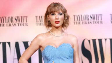 Taylor Swift stands against a background bearing her name on the red carpet of her concert film world premiere. She wears a sleeveless blue dress with a floral cut-out design, a pearl necklace and silver earrings and rings. She has her hair cut short, framing her face, and wears red lipstick and blue eyeshadow.