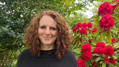 Maggie O'Farrell with curly auburn hair in black jumper. Background shows leaves on left and red flowers on the right