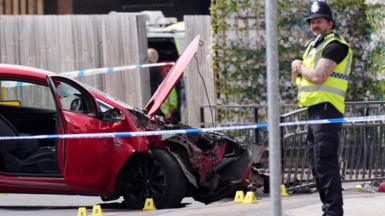 A red car with significant damage to its front is pictured behind police cordon tape. A police officers stands to the right.