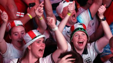 England fans dressed in red and white hats celebrate with their arms raised during a World Cup match. 