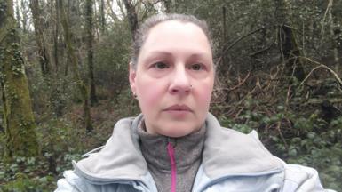 A woman with grey hair is stood centre frame. She has a solemn impression on her face and is wearing a grey turtle neck and light blue coat. She is stood in the woods.