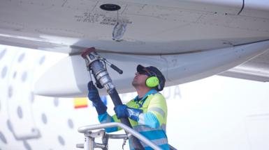 A person wearing hi vis and ear defenders lifts a jet refueling pump towards the fuel port of the wing of an aircraft on a sunny day at Seville airport in 2022.