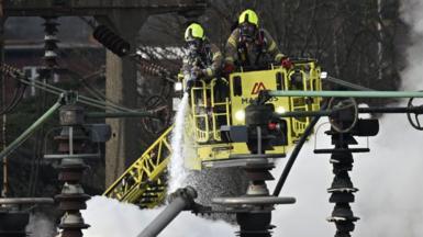 Two fire fighters wearing breathing apparatus are on board a platform which has lifted them above the electrical substation which caught fire in March. One is holding a hose which is dampening down the flames. Smoke is billowing.
