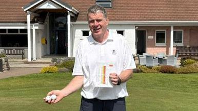 Douglas Spencer is standing outside the clubhouse with two golf balls in one hand and his score card in the other. He is wearing a white short sleeved shirt and black trousers.
