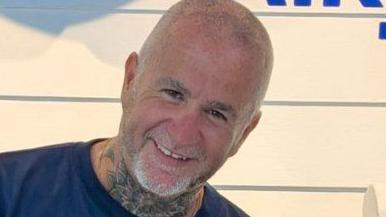 Simon Beston smiling at the camera. He has very short shaved grey hair and beard. He is wearing a blue t-shirt and has a neck tattoo. 