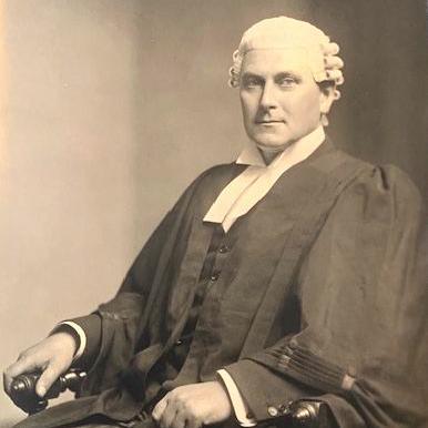 A black-and-white photo of a man dressed in a formal court gown and wig. He is sitting in a chair and looking towards the camera.