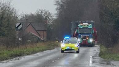 A yellow and white police car, with blue lights shining, driving along the middle of a road in front of a blue and green lorry