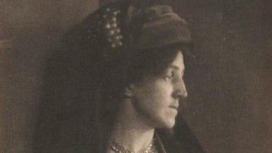 A lack and white image of a woman - May Morris - with her head turned to to left-hand side. She is wearing a head piece with long material at the back. 