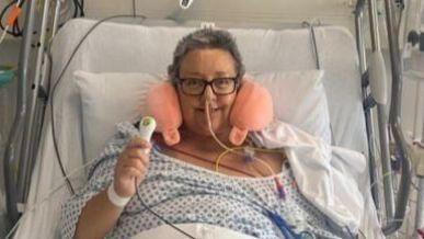 Victoria is in a hospital bed, surrounded by wires, drips and tubes. She has an inflatable neck cushion and is sat up smiling after her surgery.