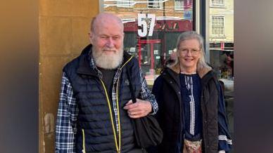 Peter on the right of this image is shown after a recent hair cut back in the UK. His beard is trimmed and his hair shorter. His wife, Barbie, stands smiling next to him.