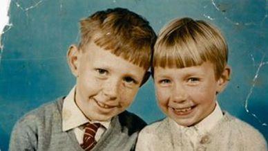 Ean Coutts as a child with one of his sisters. He has a "bowl" haircut and is smiling at the camera with his head leaning into his sister who has a similar haircut. He is wearing a school uniform - grey jumper, white shirt and maroon tie with gold stripes. His sister is also smiling and is wearing a pale cardigan over a blouse. there is a light blue background. 