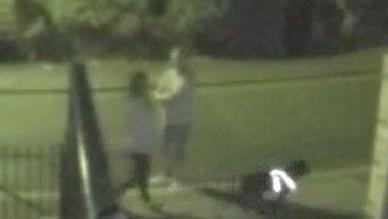 A grainy CCTV image of a dark, residential street.  A woman is walking a black dog in a reflective harness, and a man in a t-shirt and shorts appears to be turning towards her to speak to her. 