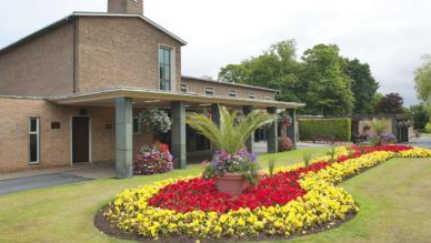 A large, colourful flowerbed with red and yellow flowers is outside the building outside the entrance. The entrance is partially covered with five columns supporting a flat roof attached to the main brick building.
