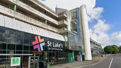 The front of St Luke's city centre pop up shop on Western Approach, Plymouth. There is a multi-storey car park above the shop. In front there is a pavement and a road on the right.