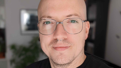 Adam Birch smiles at the camera, taking a selfie. He has wire-rimmed glasses, a pierced nose and is bald. He's wearing a black t-shirt.