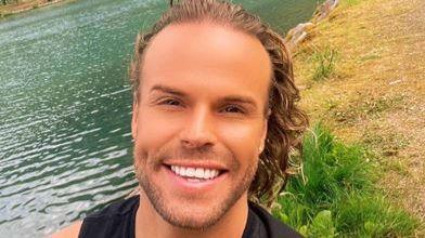 A smiling Olly Marmon looks directly at the camera, in front of a body of water and a grass bank