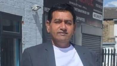 A man with black hair wearing a grey suit over a white T-shirt looks at the camera.