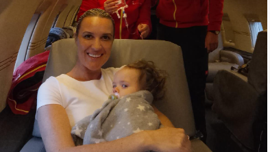 Smiling Natasha cradles her baby daughter Sienna-Rose in a grey blanket with white stars, as she sits on the plane