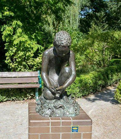 The "Child in the Park" statue in situ in Lake Meadows, sitting on a brick plinth. It is a figure of a child crouching. A bench and greenery can be seen behind it.