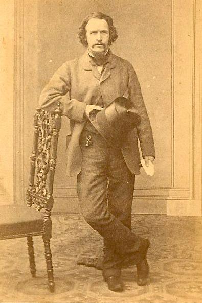 An old picture of James Smith. He is pictured standing up, leaning against a chair. He is holding a hat and wearing a suit. He has dark hair and a moustache.  