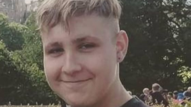 A young man with blond hair, wearing a dark grey t-shirt, smiles at the camera in a close-up shot with trees and greenery in the background. 