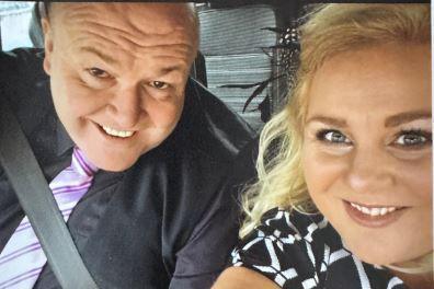 A man and woman in a car take a selfie together. The man is on the left and smiles. He is in a car as he has a seatbelt on. He has a black shirt, waistcoat and purple tie. A woman is next to him, she has blonde hair and a black and white dress on. She also smiles. It is a head and shoulders shot of the two of them.