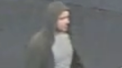 A low resolution CCTV image of a man in a grey T-shirt and brown hoodie with the hood up