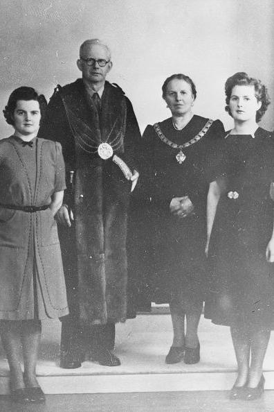 A black and white photograph showing a young Margaret Thatcher stood in a line with her parents and sister.  Her father and mother are wearing chains of office.  