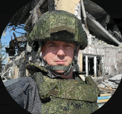 A man wearing military gear in front of a bombed-out building