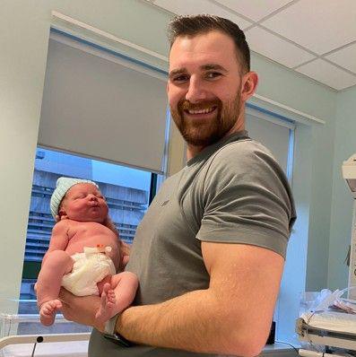 Angus Mason who is in a grey T-shirt, holds a newborn baby in a hospital room