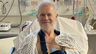 Graham Sharpe in hospital. He wears a hospital gown and one of his arms is in a navy blue sling. He has grey hair and a grey beard.