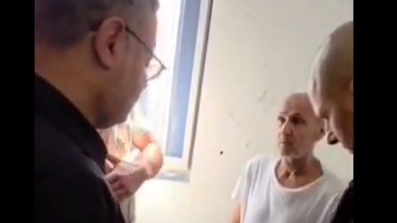 Itamar Ben Gvir, wearing glasses and a dark shirt, is seen talking to Marwan Barghouti, wearing a white shirt, who appears gaunt and aged. A police officer stands next to them with his head down. Sunlight pours through a window into the room, the walls painted off-white.